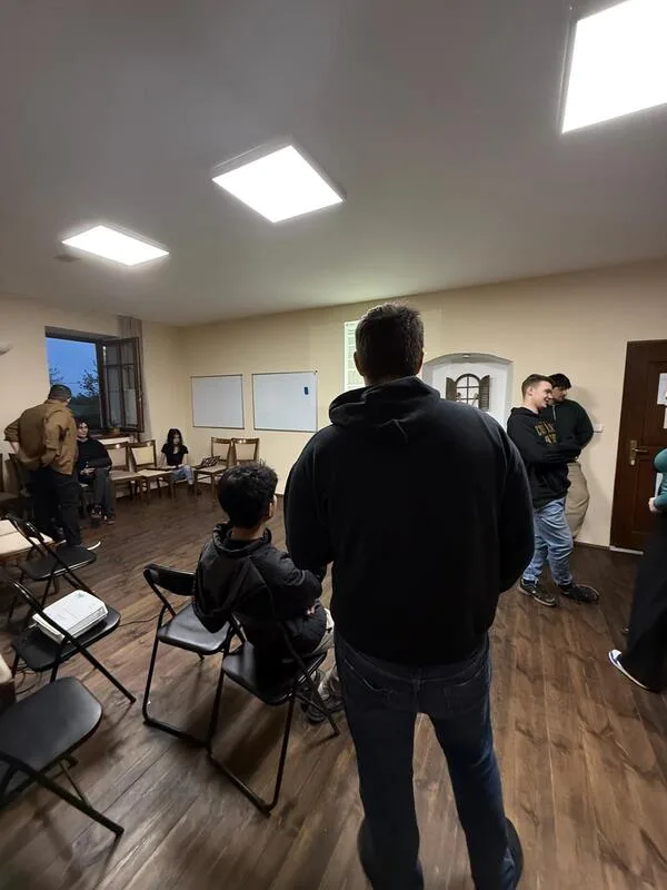 Perspective of the room of the event. Young people sitting in chairs and standing, the back of a young man in the center.