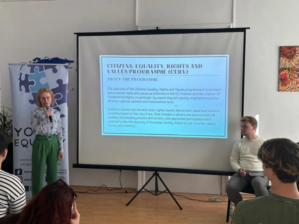 Workshop presenter talking, standing next to a projected slide, with four sitting audience members listening.